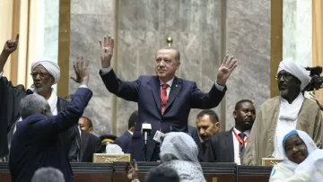 L'image montre un homme debout au centre, levant les mains en signe de salutation ou de triomphe. Il est entouré d'une foule de personnes qui semblent célébrer ou applaudir. Le décor suggère un cadre formel, probablement une assemblée ou un événement officiel, avec des personnes en habits traditionnels et des caméras visibles en arrière-plan. L'atmosphère semble être celle d'une occasion importante, marquée par une interaction entre le orateur et le public.