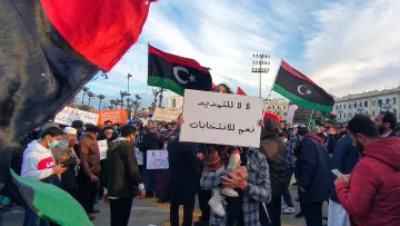 L'image montre une manifestation avec de nombreuses personnes rassemblées. Des drapeaux de la Libye flottent dans l'air, affichant des couleurs rouge, noir et vert. Au centre de la scène, une personne tient une pancarte avec des inscriptions en arabe. On peut également voir des participants portant des masques et d'autres pancartes, exprimant des revendications politiques. L'ambiance paraît dynamique, avec une multitude de personnes engagées dans une cause commune.