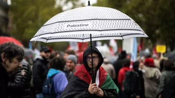 L'image montre une personne portant un parapluie noir et blanc sur lequel est écrit "Palestine". Cette personne est habillée d'un manteau sombre et se tient au milieu d'une foule qui semble participer à une manifestation. En arrière-plan, on peut apercevoir d'autres manifestants, certains portant des drapeaux et des pancartes. L'ambiance semble sérieuse et déterminée, avec des arbres et un ciel nuageux en arrière-plan.