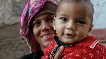 L'image montre une femme souriante tenant un bébé dans ses bras. La femme porte un foulard coloré sur la tête et a une expression chaleureuse. Le bébé, emmitouflé dans un pull rouge avec des motifs, affiche un sourire joyeux. En arrière-plan, on peut voir des murs en pierre avec un décor rustique, ce qui donne une ambiance conviviale à la scène.