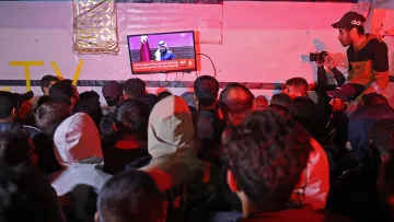 در تصویر، گروهی از افراد در حال تماشای تلویزیونی هستند که بر روی دیواری نصب شده است. تلویزیون یک برنامه خبری را نمایش می‌دهد و یک سخنران در حال صحبت است. افراد به سمت صفحه تلویزیون خیره شده‌اند و جو عمومی تصویر نشان‌دهنده توجه و کنجکاوی آن‌هاست. فضای اطراف کمی تاریک و نور محیطی کم است، به طوری که نور تلویزیون بیشتر مشخص است. برخی از افراد در لباس‌های گرم، نظیر هودی و کلاه، حضور دارند. یکی از افراد در سمت راست با دوربین در حال عکاسی است.