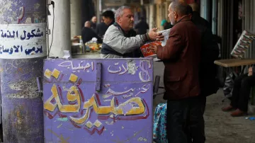 L'image montre un commerce dans une rue animée. On y voit un homme en train de discuter avec un autre, probablement en train d'échanger de l'argent ou des informations. En arrière-plan, des personnes sont assises à des tables, et on peut voir des étals remplis de marchandises. Le mur est décoré de graffitis et d'inscriptions en arabe, ainsi qu'une mention en anglais "EXCHANGE", ce qui indique probablement qu'il s'agit d'un bureau de change. L'ambiance semble vivante et typique d'un marché ou d'un bazar.