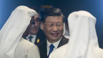 The image depicts a smiling man in a suit engaging in conversation with two individuals wearing traditional white Middle Eastern attire, including headscarves. The setting feels formal and diplomatic.