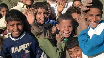 L'image montre un groupe d'enfants qui sourient joyeusement. Ils semblent apprécier le moment, avec des expressions de joie et beaucoup d'énergie. Les enfants sont vêtus de vêtements variés et certains lèvent la main ou se penchent vers l'avant, montrant un sentiment d'excitation et d'unité. L'arrière-plan est flou, mettant en avant leurs visages animés et heureux.