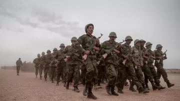 L'image représente un groupe de soldats marchant en formation sur un chemin poussiéreux. Ils sont habillés en tenue militaire camouflée et portent des casques. Chaque soldat tient un fusil à la main. L'atmosphère semble sérieuse, et le ciel est nuageux, suggérant peut-être des conditions climatiques difficiles. En arrière-plan, on aperçoit une ligne de végétation ou d'arbres, ajoutant à l'impression d'un environnement militaire.