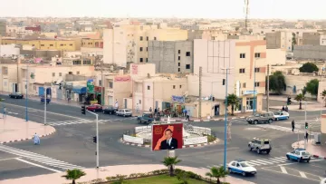 L'image montre une vue urbaine d'une ville. On y aperçoit des bâtiments de plusieurs étages, avec une architecture simple. À l'avant, il y a un rond-point avec un grand panneau affichant un portrait d'une figure publique. Plusieurs voitures sont stationnées ou circulent autour du rond-point. Des arbres et des éléments de signalisation routière ajoutent à l'ambiance de la scène. Le ciel est clair, suggérant une atmosphère calme, et la lumière semble indiquer une période de la journée avancée.