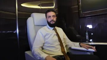 Un homme est assis dans un fauteuil confortable à l'intérieur d'un avion privé. Il porte une chemise claire et une cravate. L'environnement est élégant, avec des surfaces sombres et brillantes.