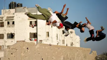 La imagen muestra a un grupo de jóvenes realizando acrobacias o saltos, en un entorno urbano. Están en el aire, ejecutando movimientos como un salto hacia atrás, mientras que de fondo se pueden ver edificios con paredes desgastadas. Los chicos parecen estar disfrutando de la actividad, mostrando energía y alegría. La escena refleja un momento de juego y diversión en un área urbana.