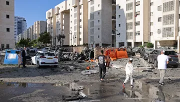 L'immagine mostra una scena di distruzione in un'area urbana. Si possono vedere edifici residenziali con facciate chiare, circondati da macerie e detriti. Alcune auto sono danneggiate, con pezzi sparsi sul terreno. Ci sono persone che camminano nella strada, apparentemente mentre osservano il danno. Il cielo è sereno, e ci sono anche pozzanghere d'acqua, suggesting che recente pioggia potrebbe essere rientrata nell'area. La scena trasmette un senso di caos e di recente violenza o attacco.