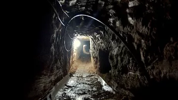 L'image montre un tunnel sombre creusé dans la terre. Les parois de la cavité sont en pierre, et le sol est inégal, probablement en terre battue. Une lumière blanche éclaire partiellement le tunnel, révélant son intérieur. Le fond du tunnel semble s'étendre à l'infini, créant une ambiance mystérieuse. Des câbles électriques sont visibles à certains endroits, indiquant qu'il pourrait y avoir des installations techniques à l'intérieur.