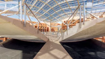 L'image montre un intérieur architectural moderne avec une structure impressionnante. Le plafond est en verre, permettant à la lumière naturelle d'entrer, et crée une ambiance lumineuse. Les murs et les surfaces sont en béton et en bois, offrant un contraste intéressant. Des passerelles en bois traversent l'espace, menant à des zones ouvertes. La conception suggère un espace fonctionnel et esthétique, propice à des activités variées.