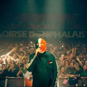 Un artiste en concert, masqué, devant une foule enthousiaste, avec des drapeaux irlandais et palestiniens.