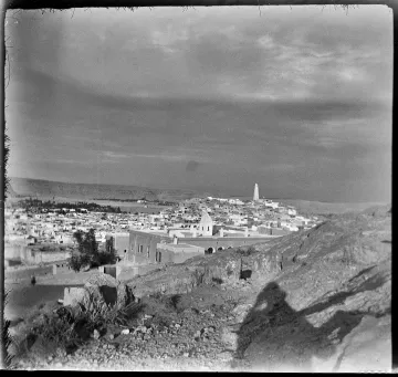 L'image montre un paysage urbain, probablement d'une ville historique. On peut voir des maisons blanches et des structures architecturales qui s'étendent à travers la vallée. À l'arrière-plan, un minaret s'élève, suggérant une présence religieuse. Le ciel semble nuageux, ce qui ajoute une ambiance dramatique à la scène. Le terrain est rocailleux au premier plan, et l'éclairage donne une impression de nostalgie, typique des photographies anciennes.