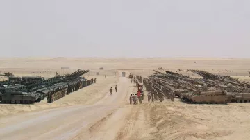 The image depicts a vast desert landscape, characterized by a flat, sandy terrain. On either side of a wide dirt road, there are rows of military tanks and armored vehicles arranged in a formation. Several soldiers are visible on the road, possibly coordinating or observing the vehicles. The background features an expansive desert with minimal vegetation, emphasizing the military setup in a stark and barren environment. The scene conveys a strong military presence and readiness.