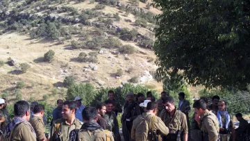 الصورة تظهر مجموعة من الرجال يرتدون ملابس عسكرية في منطقة طبيعية. توجد تلال خضراء في الخلفية، وبدت بعض الأشجار. يبدو أن هؤلاء الرجال يتفاعلون مع بعضهم البعض، حيث هناك من يتحدث ومن يتواجد في مجموعات صغيرة. يظهر في الصورة أيضا بعض الأشخاص يحملون أسلحة. الجو العام يوحي بنشاط اجتماعي أو تحضير لشيء ما.