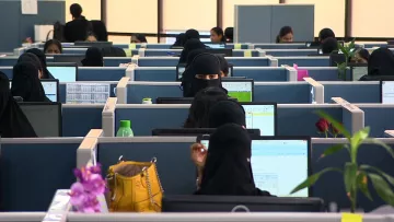 L'image montre un espace de bureau moderne, avec plusieurs employés assis à leurs bureaux, chacun devant un ordinateur. La plupart des femmes portent des vêtements noirs, y compris des hijabs ou des niqabs, ce qui leur donne une apparence uniforme. On peut voir des écrans d'ordinateur affichant des tableaux ou des données, ainsi que quelques plantes décoratives et des objets sur les bureaux. L'ambiance semble professionnelle et organisée, avec un grand nombre de personnes travaillant dans cet environnement administratif.