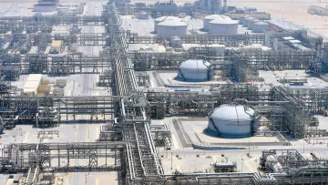 L'immagine mostra un impianto industriale, probabilmente una raffineria o un complesso di estrazione di prodotti petroliferi. Si possono vedere grandi serbatoi rotondi e una vasta rete di tubazioni che si snodano attraverso il sito. L'area circostante appare deserta, con una superficie di sabbia o terra arida. Sullo sfondo si intravedono strutture alte, che potrebbero essere torri di raffineria o impianti di trattamento. L'intero complesso è ben organizzato, con una disposizione sistematica delle tubazioni e dei serbatoi.