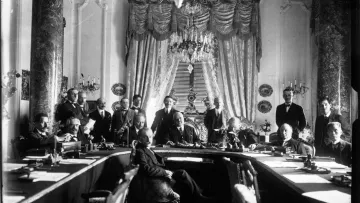 L'image montre une grande réunion dans une salle ornée. Au centre, plusieurs hommes sont assis autour d'une grande table, tandis que d'autres se tiennent debout en arrière-plan. La pièce est décorée avec des rideaux élégants et des éléments architecturaux raffinés, tels que de grands lustres et des colonnes. Les participants semblent engagés dans une discussion sérieuse, reflétant l'importance de l'événement. L'atmosphère est formelle et historique.