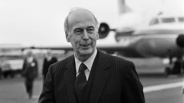 L'image montre un homme en costume rayé, marchant sur une piste d'aéroport. Il affiche un léger sourire et semble détendu. En arrière-plan, on peut apercevoir un avion et d'autres personnes qui se déplacent. L'ambiance générale est celle d'un moment de rencontre ou d'accueil, et la photographie est en noir et blanc, ce qui lui donne un caractère classique.
