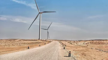 La imagen muestra un paisaje árido y desértico con un camino de grava que se extiende hacia el horizonte. A ambos lados del camino, se pueden ver aerogeneradores, que son grandes turbinas eólicas que aprovechan la energía del viento. El cielo está despejado y presenta un tono azul, lo que sugiere un día soleado. La escena transmite una sensación de tranquilidad y sostenibilidad, con la tecnología eólica destacando en un entorno natural.