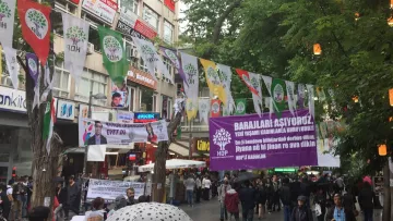 L'image montre une rue animée avec des banderoles accrochées entre les bâtiments. Ces banderoles semblent promouvoir un événement ou une cause, avec des slogans en caractères visibles. On peut également voir des groupes de personnes se rassembler au sol, suggérant une atmosphère de manifestation ou de réunion. Des arbres bordent la rue, ajoutant une touche de verdure à la scène. La journée semble nuageuse, ce qui pourrait indiquer un temps frais.