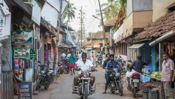 L'image montre une rue animée d'un village ou d'une petite ville. On y voit des bâtiments aux façades colorées et typiques de l'architecture locale, avec des toits en tuiles. Plusieurs personnes sont présentes, certaines se déplaçant à pied, d'autres sur des motos. Des échoppes bordent la rue, vendant probablement des fruits et légumes. L'atmosphère semble vivante et animée, avec des fils électriques et des palmiers visibles en arrière-plan.