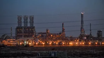 La imagen muestra una instalación industrial, probablemente una refinería o planta petroquímica, iluminada al anochecer. Se pueden ver numerosas estructuras metálicas altas, chimeneas y torres, con luces brillantes que resaltan en el entorno oscuro. Hay líneas eléctricas que cruzan la imagen y el paisaje es árido, lo que sugiere que la planta está ubicada en una zona desértica o poco vegetada. La atmósfera transmite una sensación de actividad industrial, con un enfoque en la tecnología y la producción.