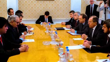 The image depicts a formal meeting taking place around a long wooden table in what appears to be a conference room. Participants are seated on both sides of the table, engaged in conversation or listening attentively. The table is set with various documents, and there are snacks and beverages in front of some attendees. The room has an elegant decor with a patterned wall and paintings. A few individuals are standing in the background, likely observing the meeting. The atmosphere seems serious and focused on discussion.