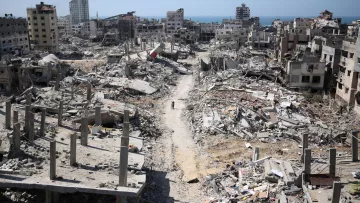 L'immagine mostra un paesaggio di devastazione, con edifici in rovina e macerie sparse in tutta l'area. Le strutture, una volta abitate, sono ridotte a scheletri di cemento e detriti. In lontananza, si scorge il mare, che contrasta con la desolazione del terreno. Una persona si muove tra le macerie, evidenziando la vastità della distruzione. Il cielo è sereno, creando un contrasto inquietante con la scena di desolazione sottostante.