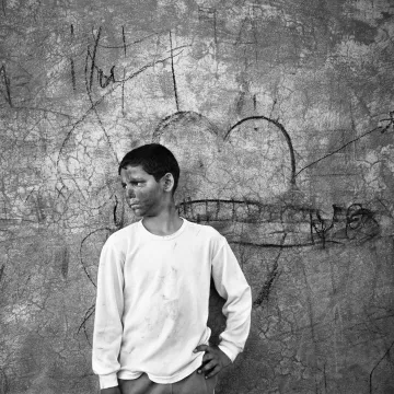 L'image montre un garçon qui se tient contre un mur texturé, où l'on peut voir des éraflures et des graffitis. Il porte un t-shirt blanc légèrement sale et un pantalon beige. Son expression semble pensive ou mélancolique. Le décor derrière lui, avec un dessin de cœur, contraste avec son attitude, créant une atmosphère intéressante. Cela donne une impression de nostalgie ou de réflexion sur son environnement.