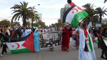 L'image montre une manifestation avec des personnes marchant dans une rue, portant des drapeaux représentant le Sahara occidental. Il y a une atmosphère festive, avec des participants qui tiennent des banderoles et des ballons. Des palmiers se dressent en arrière-plan, ajoutant à l'ambiance. Les manifestants semblent exprimer leurs revendications et leur solidarité.