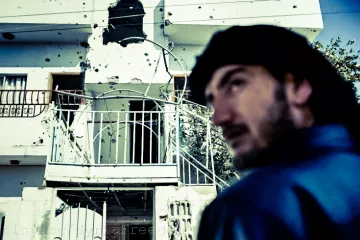 L'image montre un bâtiment portant des marques visibles d'impacts de balles, suggérant des dommages dus à des conflits. Au premier plan, un homme est partiellement visible, tourné vers l'extérieur, avec une expression pensive. L'environnement semble urbain, et le contraste entre le visage de l'homme et le bâtiment endommagé crée une atmosphère de tension et de tristesse.