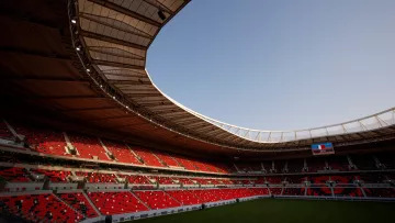 L'image montre un stade moderne avec des sièges rouges et une large toiture. La pelouse est bien entretenue, et on peut apercevoir un écran géant à une extrémité. Le ciel est dégagé, indiquant une belle journée. L'intérieur du stade semble prêt à accueillir un événement, avec une atmosphère calme et anticipative.