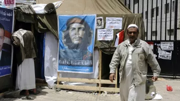 L'image montre un campement avec une tente, devant laquelle se trouve une affiche représentant un homme avec un béret, souvent associé à Che Guevara. À côté de l'affiche, on peut voir des affiches supplémentaires et une personne marchant, vêtue d'une tenue clair. L'ambiance semble refléter un contexte politique ou social, probablement avec des éléments de protestation ou de revendication.