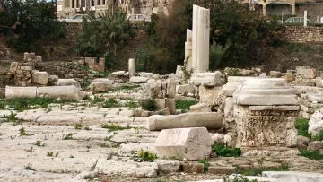 L'image montre un site archéologique avec des ruines anciennes. On peut voir des colonnes et des blocs de pierre, certains sont en désordre, tandis que d'autres semblent être en place. En arrière-plan, il y a des bâtiments plus récents, probablement des habitations, qui contrastent avec les vestiges historiques. La végétation est présente autour des pierres, avec des herbes et des fleurs qui poussent dans les fissures. L'ensemble évoque un mélange de l'histoire ancienne et de la vie moderne.