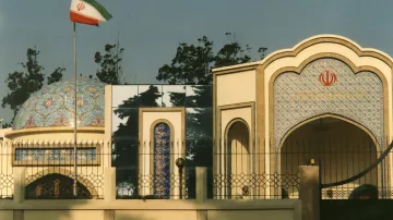 L'image montre un bâtiment architectural de style iranien, typique avec des tuiles colorées et des motifs ornementaux. On peut voir le drapeau iranien flottant sur un mât à gauche. Le bâtiment présente une structure avec un dôme décoré et des arcs, intégrant des éléments de design traditionnels. L'environnement est arboré, avec des arbres visibles en arrière-plan.