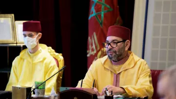 L'image montre une réunion se déroulant dans un cadre officiel. À droite, un homme vêtu d'une tenue traditionnelle marocaine de couleur jaune, portant un fez rouge, est attentif et semble concentré sur les discussions. À gauche, un jeune homme portant également un jaune et un masque facial regarde devant lui. En arrière-plan, on peut apercevoir des éléments de décoration et des drapeaux, évoquant le contexte marocain de la scène. L'ambiance générale semble sérieuse et solennelle.