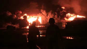 The image depicts two figures walking away from a large fire at night, with bright flames and thick smoke rising into the sky. The scene conveys a sense of urgency and danger.