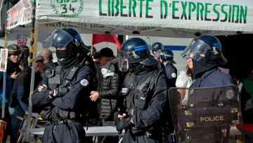 L'image montre un groupe de policiers en uniforme, équipés de casques et de protections, se tenant devant un stand ou une tente avec l'inscription "LIBERTE D'EXPRESSION". On peut voir plusieurs personnes en arrière-plan, probablement des manifestants ou des observateurs, dans un environnement urbain. L'atmosphère semble être celle d'un rassemblement ou d'une manifestation, avec des thèmes liés à la liberté d'expression.