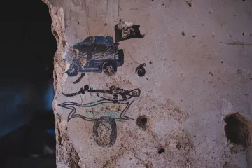 L'image montre un mur en béton décrépit où des graffitis sont visibles. On peut y voir une représentation d'un véhicule, probablement une voiture de police, accompagnée d'un drapeau. En dessous, il y a un dessin stylisé d'un squelette, surmonté d'une forme qui pourrait évoquer un traîneau ou un chariot. Le mur lui-même présente des traces d'usure et des fissures, ajoutant une dimension d'abandon à la scène.