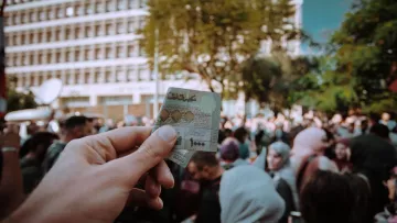 در این تصویر، یک دست در حال نگه‌داشتن یک کارت شناسایی یا مدرک مشابهی است. در پس‌زمینه، جمعیتی از افراد دیده می‌شود که در یک مکان عمومی جمع شده‌اند. برخی از افراد چادر یا پوشش‌های اسلامی دارند. نور روز و فضای خارجی نشان‌دهنده یک رویداد یا تجمع عمومی است. احساس عمومی شلوغی و فعالیت اجتماعی در این تصویر مشهود است.