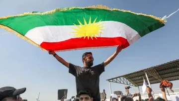 در این تصویر، فردی را مشاهده می‌کنیم که در بالای شانه‌های یک شخص دیگر ایستاده و پرچم کردستان را به اهتزاز درآورده است. پرچم دارای رنگ‌های سبز، سفید و قرمز با خورشید زرد در وسط است. جمعیتی در اطراف آنها حضور دارد و به نظر می‌رسد در حال برگزاری یک تظاهرات یا تجمع هستند. فضای تصویر آکنده از انرژی و احساسات جمعی است.