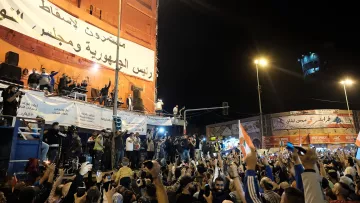 L'image montre une grande foule rassemblée dans une place publique, manifestant avec enthousiasme. Les gens lèvent les mains et brandissent des drapeaux, exprimant des émotions fortes. À l'arrière-plan, on peut voir un grand panneau affichant des messages politiques. Des personnages sont visibles sur une scène surélevée, semblant s'adresser à la foule. L'ambiance semble festive et dynamique, avec des lumières qui illuminent la scène.