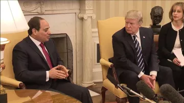 L'image montre deux hommes assis dans un salon. À gauche, un homme en costume sombre et cravate rouge parle avec une expression attentive. À droite, un homme en costume sombre et cravate rayée semble écouter, avec une expression concentrée. Ils sont entourés de mobilier élégant et d'une ambiance formelle, indiquant une réunion importante. À l'arrière-plan, on peut voir une cheminée et une sculpture. Une femme est également présente, mais sa position et son rôle ne sont pas clairement indiqués.