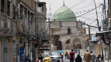 L'image montre une rue animée dans une ville. On y voit des bâtiments anciens avec des façades en pierre, certains étant en cours de rénovation. Au fond, une grande coupole verte domine la scène, indiquant la présence d'un bâtiment religieux. Les fils électriques sont visibles au-dessus de la rue. Des personnes marchent sur le trottoir, et une moto ainsi qu'une voiture jaune circulent. L'atmosphère semble à la fois traditionnelle et dynamique.