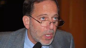Je ne sais pas qui est cette personne. Cependant, je peux dire que l'image semble montrer un homme portant des lunettes, avec une barbe grisonnante et des cheveux également grisonnants. Son expression semble sérieuse et il est peut-être en train de parler ou de faire une présentation. Son veston est en tissu à motifs.