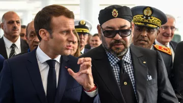 L'image montre deux hommes en discussion, entourés de plusieurs personnes. L'un porte un costume sombre et une cravate, tandis que l'autre est habillé d'une manière plus formelle, avec des lunettes de soleil et un turban. Ils semblent engagés dans une conversation sérieuse, et l'environnement semble être une cérémonie officielle ou un événement important. Les expressions sur leurs visages suggèrent qu'ils sont en train de partager des idées ou des informations.