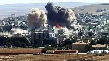 L'image montre un paysage urbain après une explosion, avec d'importants nuages de fumée et des décombres. Des bâtiments sont partiellement détruits, et l'atmosphère est sombre en raison de la fumée épaisse. Le sol semble être rural, avec des champs et quelques structures en arrière-plan. Cette scène évoque des conséquences dramatiques d'un conflit ou d'une attaque.