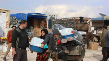 The image shows a busy scene where people are engaged in various activities, likely in a market or a distribution area. In the foreground, a man and a woman are interacting next to a small vehicle loaded with large containers, possibly for water or other supplies. The woman is dressed in traditional clothing and appears to be holding a tray, while the man seems to be dressed in casual attire. In the background, several people are seen unloading boxes and organizing goods, with a truck and some tents visible. The setting has a somewhat informal atmosphere, suggestive of a community effort related to aid or distribution.