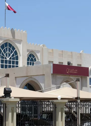 L'image montre un bâtiment de style architectural arabe moderne, avec une grande fenêtre en arc sur le mur. Au-dessus de l'entrée, il y a une enseigne en arabe et en anglais avec le nom "Qatar Charity". Le bâtiment est entouré de parasols et d'une clôture, et on peut voir un drapeau du Qatar flottant au sommet du bâtiment. Le ciel est dégagé et ensoleillé.